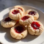 A plate of freshly baked raspberry thumbprint cookies, coated in sugar and filled with bright red jam.