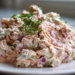 A mound of creamy salmon salad mixed with red onion and herbs, topped with fresh dill.