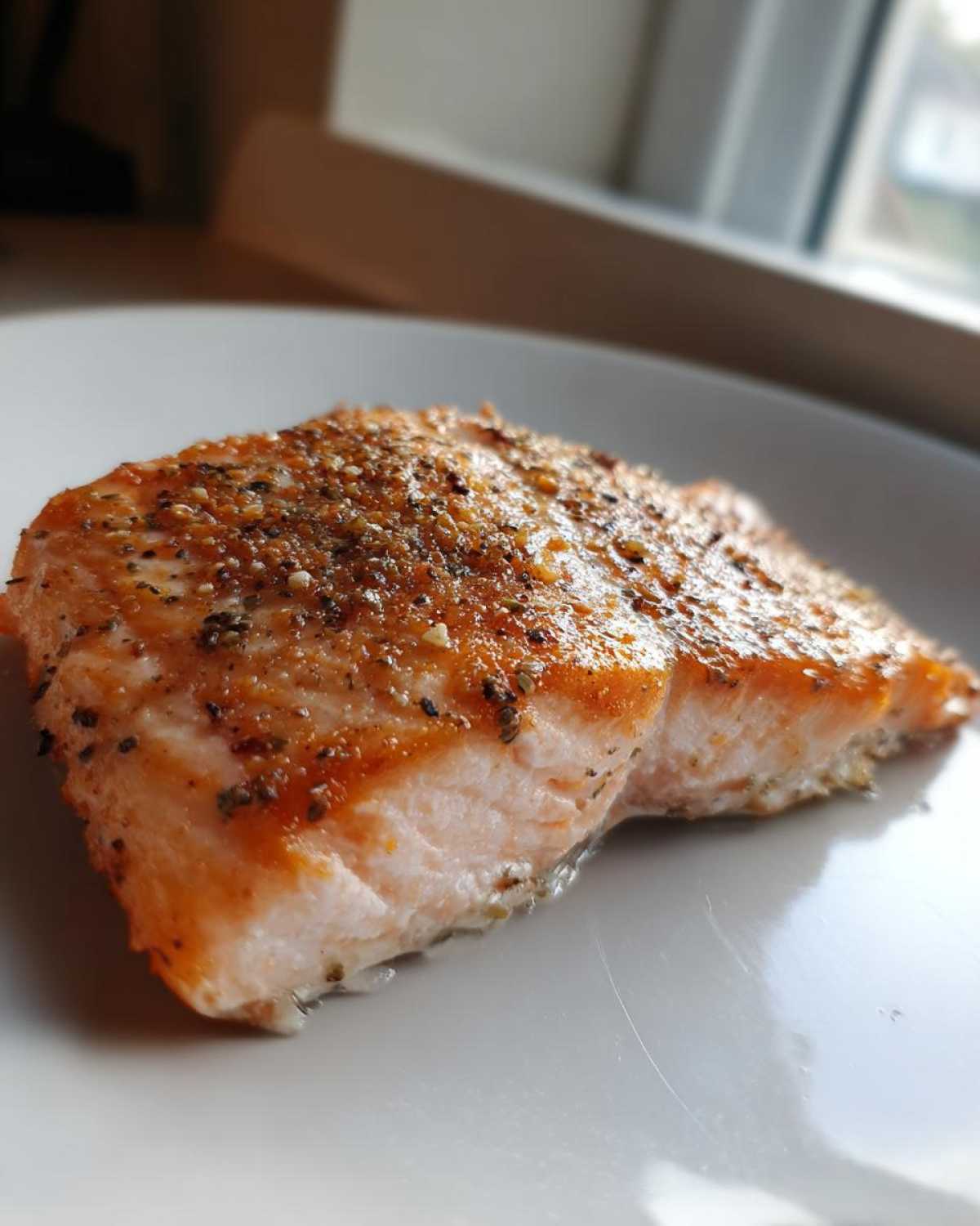 Close-up of a juicy, perfectly cooked piece of air fryer salmon seasoned with pepper and herbs on a white plate.