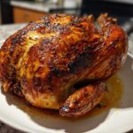 A whole roasted chicken with crispy, seasoned skin resting on a white plate, made using an air fryer whole chicken recipe.