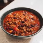 Close-up of a dark bowl filled with hearty chili, featuring ground beef, kidney beans, and rich tomato sauce.