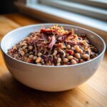 A white bowl filled with savory black eyed peas mixed with bacon and topped with shredded smoked meat.