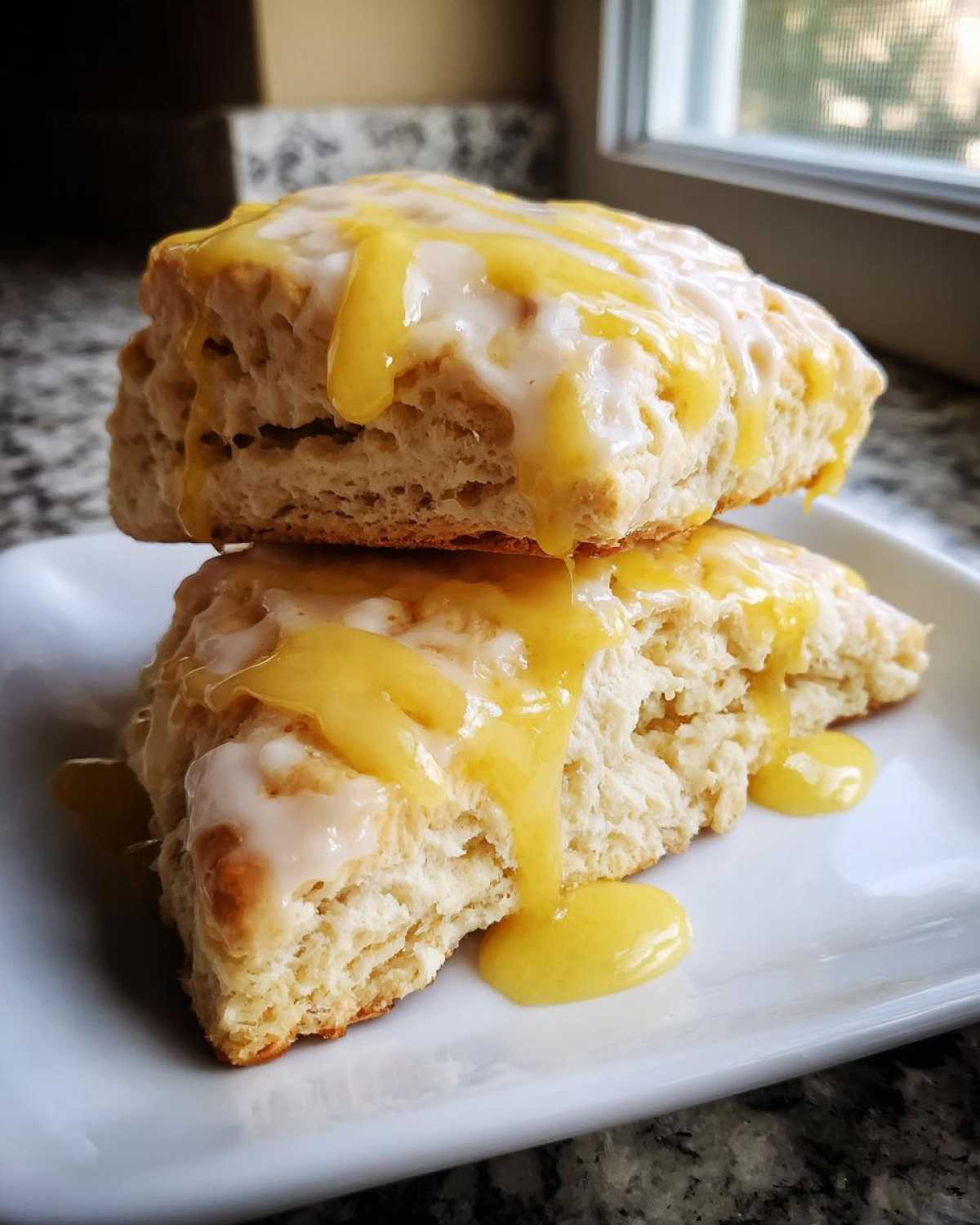 Two stacked, fluffy lemon scones topped with white and bright yellow lemon glaze dripping down the sides.