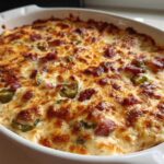 A close-up of a baked popper dip in a white oval dish, featuring melted, browned cheese, bacon pieces, and jalapeño slices.