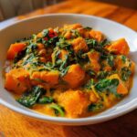 Close-up of a white bowl filled with vibrant orange pumpkin curry mixed with dark green wilted spinach.