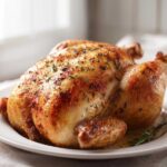 A whole, golden-brown roasted chicken seasoned with herbs resting on a white plate with pan juices.