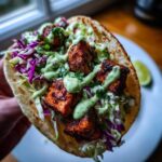 A close-up of a handheld salmon taco filled with blackened salmon chunks, purple cabbage slaw, and a creamy green sauce.