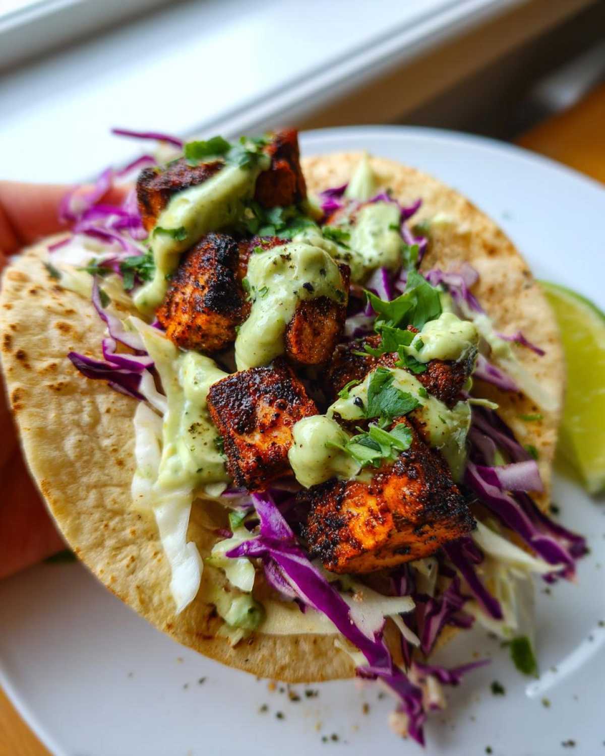 A close-up view of a single, fully loaded salmon taco featuring blackened salmon chunks, purple and white cabbage slaw, and a drizzle of green avocado crema.