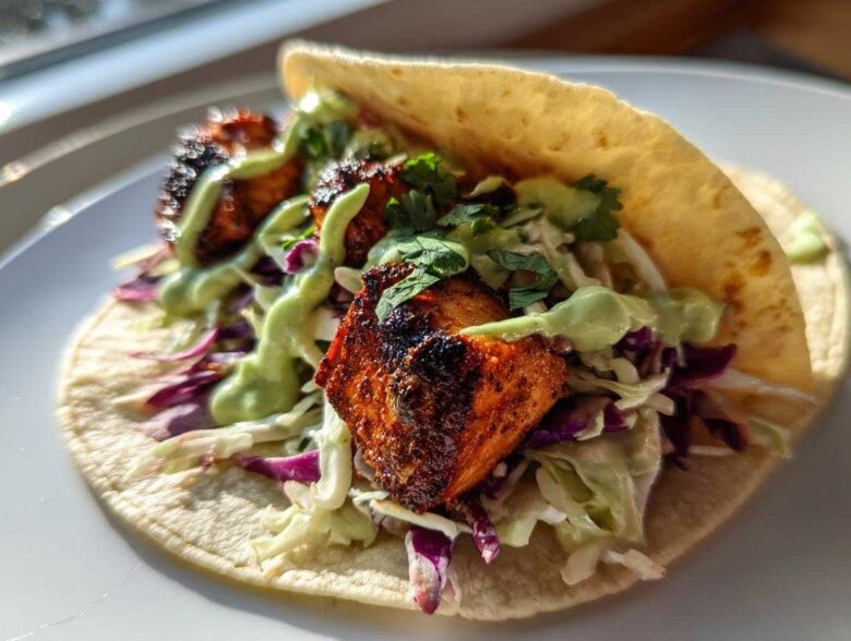 A close-up of a single, delicious salmon taco filled with blackened salmon chunks, purple cabbage slaw, and drizzled with a light green sauce.