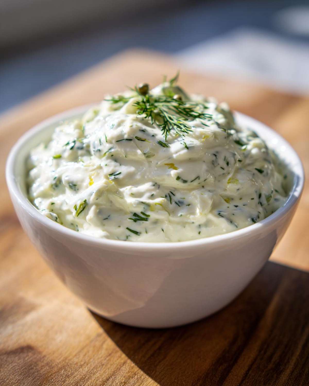 Close-up of creamy homemade tartar sauce garnished with fresh dill and a caper in a white bowl.