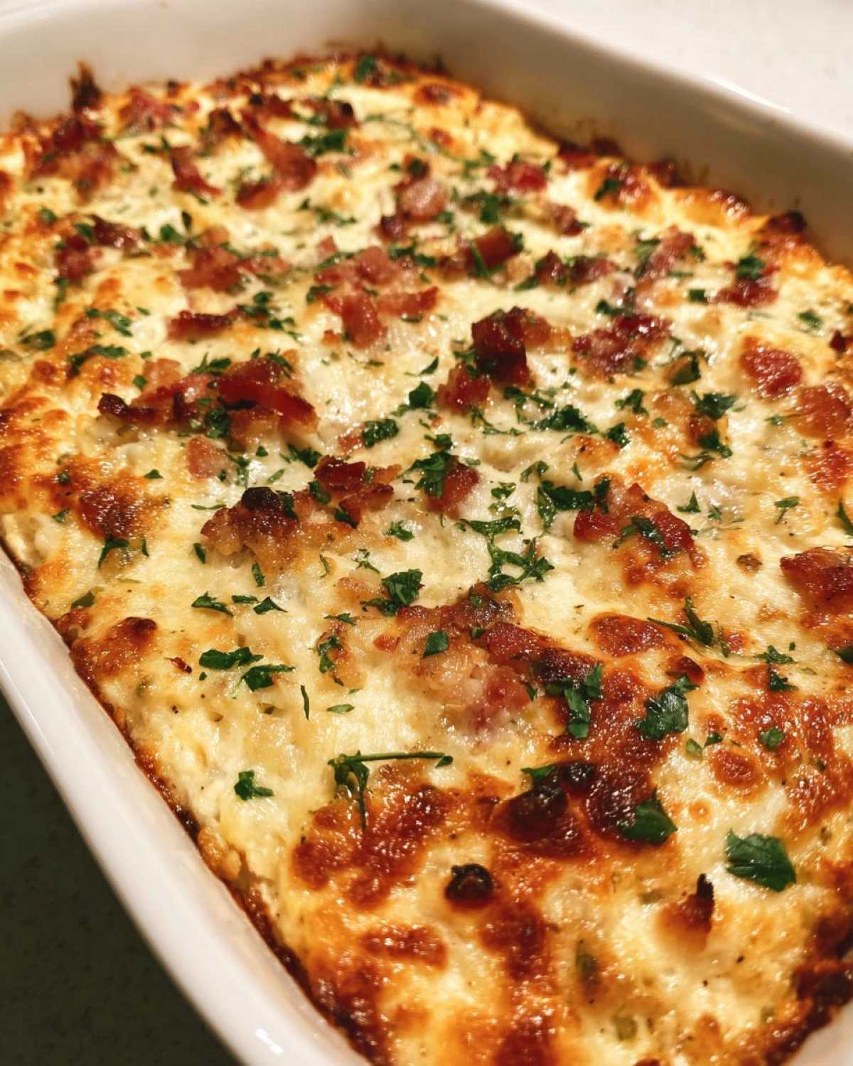 Close-up of a baked, cheesy popper dip topped with crispy bacon bits and fresh parsley in a white baking dish.