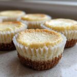 Several baked mini cheesecake bites with golden tops resting in white paper liners on a light countertop.
