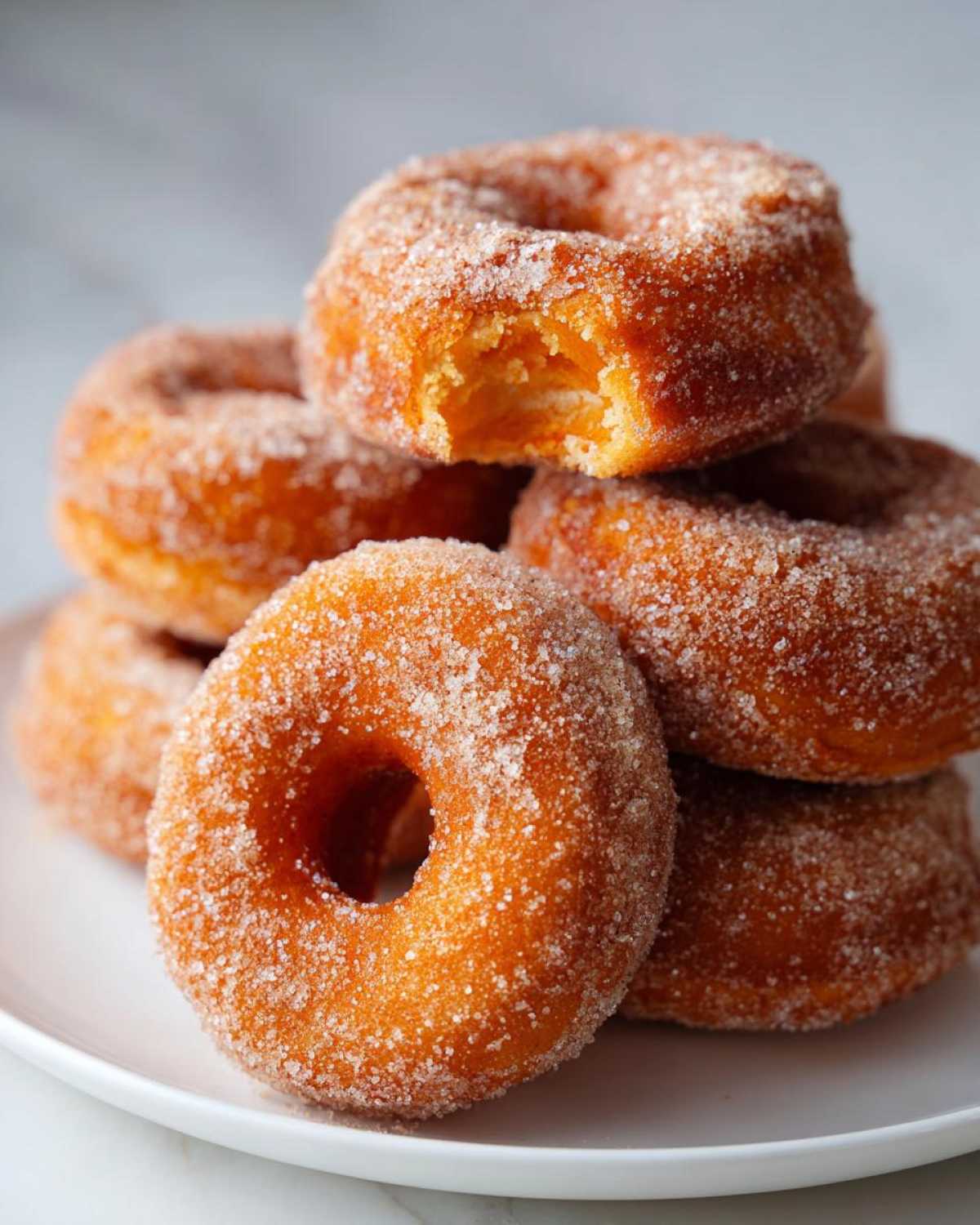 A close-up stack of golden baked pumpkin donuts heavily coated in sparkling sugar, one donut has a bite taken out.