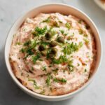 Close-up of a creamy, pale pink smoked salmon dip garnished generously with fresh dill and whole capers in a white bowl.