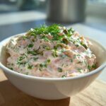 A close-up of a creamy smoked salmon dip topped with fresh dill and capers in a white bowl.