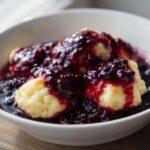 A white bowl filled with fluffy dumplings smothered in a rich, dark blackberry sauce, showcasing the texture of the Blackberry Dumplings.