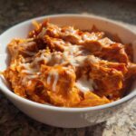A white bowl filled with shredded, orange-sauced slow cooker buffalo chicken chili topped with melted white cheese.