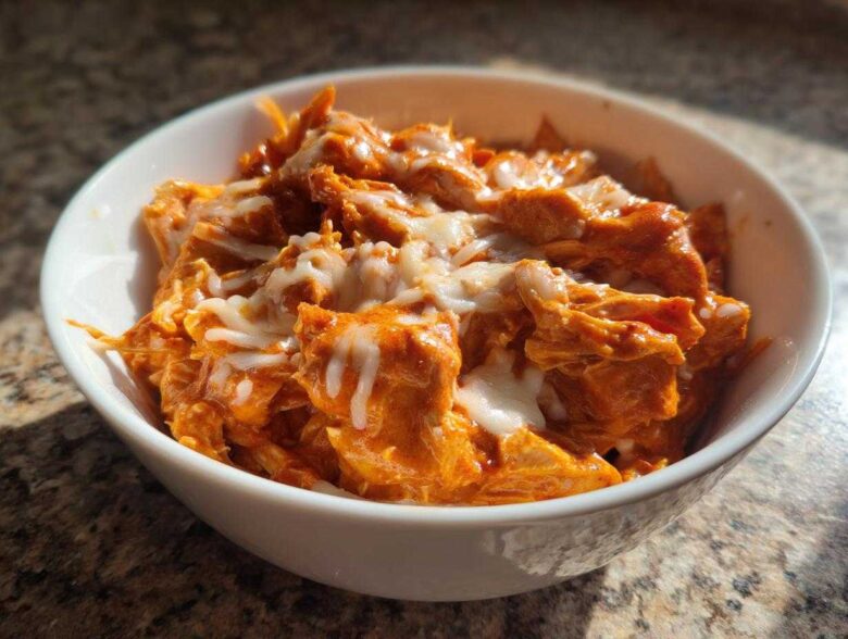 A white bowl filled with shredded, orange-sauced slow cooker buffalo chicken chili topped with melted white cheese.