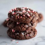 A stack of three chewy, dark chocolate peppermint chocolate chip cookies topped with crushed candy canes.