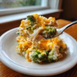 A spoonful of creamy chicken broccoli rice casserole topped with melted cheddar cheese being lifted from a white plate.
