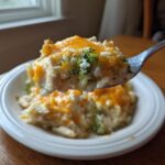 A spoonful of creamy chicken broccoli rice casserole topped with melted cheddar cheese being lifted above a white plate.