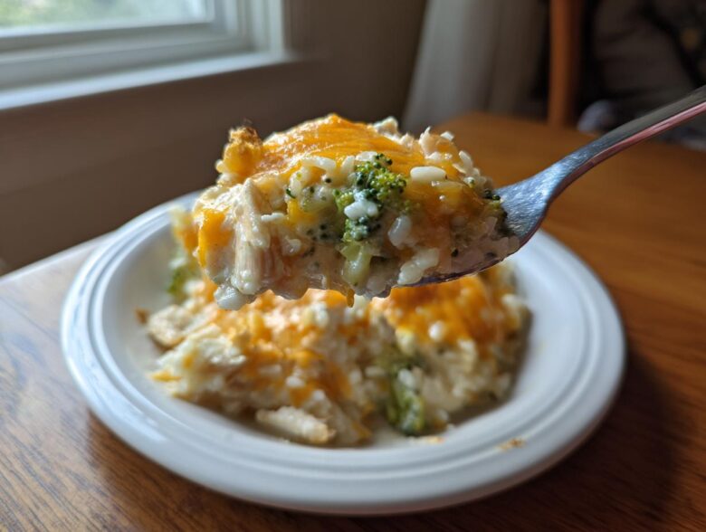 A spoonful of creamy chicken broccoli rice casserole topped with melted cheddar cheese being lifted above a white plate.