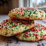 A stack of three soft, round christmas sprinkle sugar cookies topped with red, green, and white sprinkles.