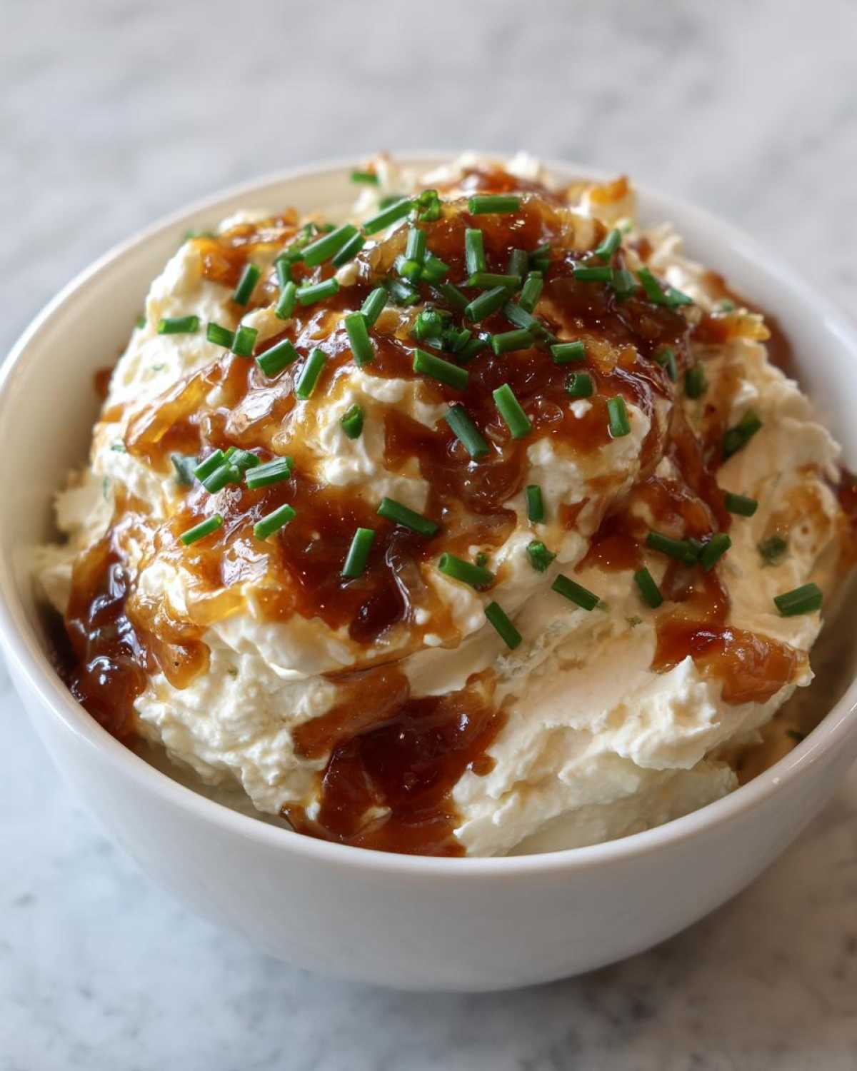 Close-up of rich, creamy caramelized onion dip topped with sweet onions and fresh green chives in a white bowl.