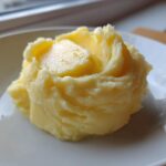 A scoop of ultra creamy mashed potatoes topped with a melting pat of butter on a white plate.