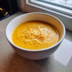 A close-up of a vibrant orange cauliflower soup topped with melted cheese and black pepper in a white bowl.