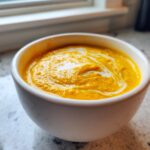Close-up of a vibrant orange cauliflower soup, swirled with cream, served in a white bowl.