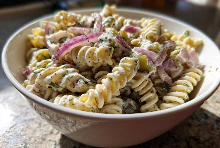 Close-up of a creamy pickle pasta salad made with rotini pasta, red onion, and dill, served in a white bowl.