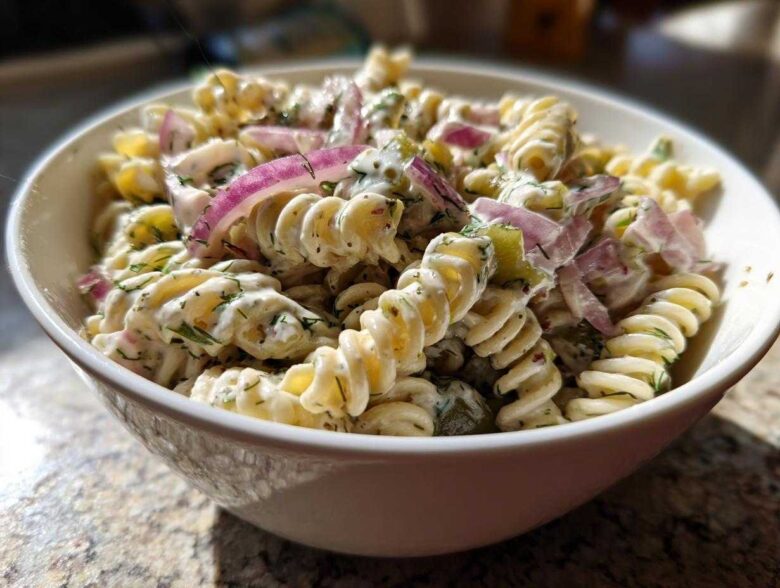 Close-up of a creamy pickle pasta salad made with rotini pasta, red onion, and dill, served in a white bowl.