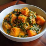 A white bowl filled with rich, orange pumpkin curry featuring large chunks of pumpkin and dark green spinach.
