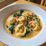 A close-up of creamy ravioli soup featuring tortellini, wilted spinach, and crumbled sausage in a rich broth.