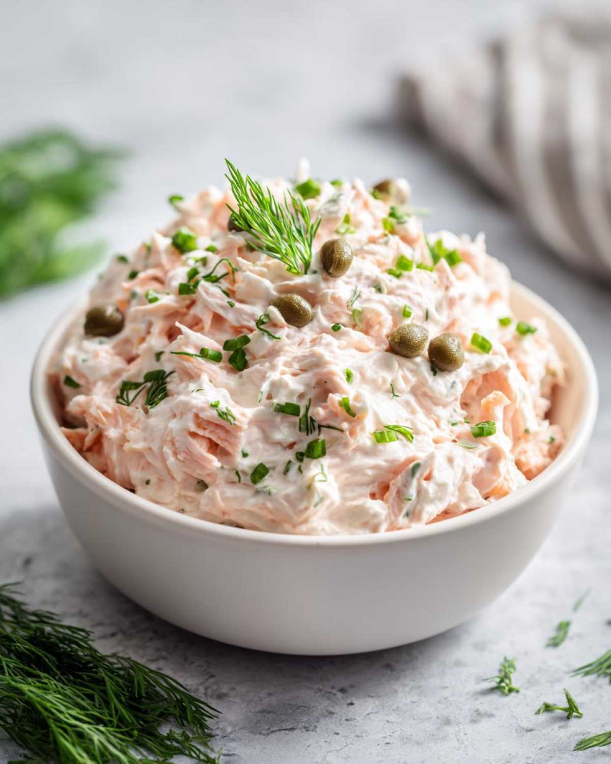 A close-up of creamy smoked salmon dip garnished with capers and fresh dill in a white bowl.