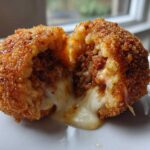 A crispy, golden-brown rice balls cut in half showing a savory meat filling and gooey melted mozzarella cheese.