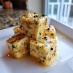 Close-up of four cubes of marinated cheese, glistening with oil and herbs, served on a white plate.