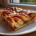 Close-up of a glistening, caramelized slice of easy apple tart resting on a white plate near a window.