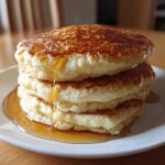 A tall stack of four fluffy almond flour pancakes drizzled generously with maple syrup.