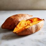 Close-up of a perfectly baked sweet potatoes, one whole and one split open revealing fluffy orange flesh.