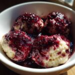 Close-up of four fluffy Blackberry Dumplings served warm in a white bowl, generously topped with dark, rich blackberry sauce.