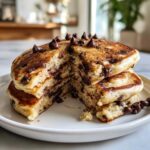 A stack of fluffy chocolate chip pancakes cut open to show melted chocolate inside, topped with whole chocolate chips.