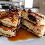 A stack of fluffy chocolate chip pancakes cut open, drizzled with syrup and topped with chocolate chips.