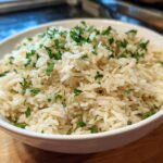 A close-up of fluffy white rice pilaf served in a white bowl and garnished with fresh chopped parsley.