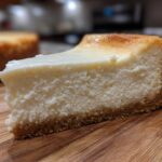 A thick, creamy slice of new york cheesecake with a golden-brown top and graham cracker crust, resting on a wooden board.
