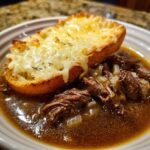 Close-up of tender shredded beef in rich broth with onions, topped by a slice of toasted bread covered in melted cheese for french onion beef stew.
