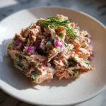 A mound of creamy salmon salad mixed with red onion and fresh dill, served on a white plate.