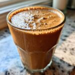 Close-up of a rich, brown Coffee Smoothie with a foamy top in a clear glass, sitting on a granite counter near a window.