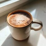 Close-up of rich mexican hot chocolate with foam in a speckled ceramic mug, lit by sunlight.